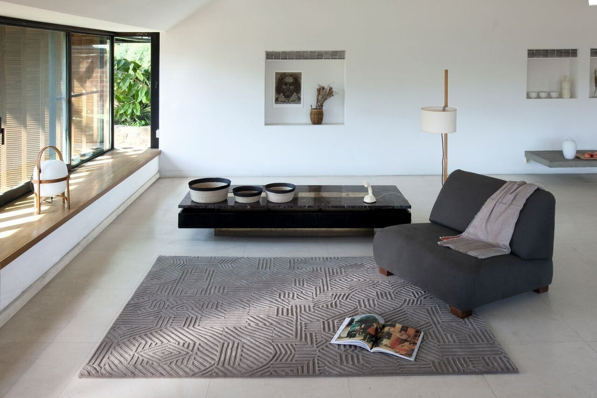 Gray upholstered chair on a patterned gray rug in a modern, minimalist living room with black coffee table and natural light.