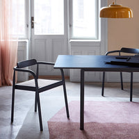Black table and chair with muted pink rug, warm yellow lamp, and soft light from large windows. Modern minimalist design.