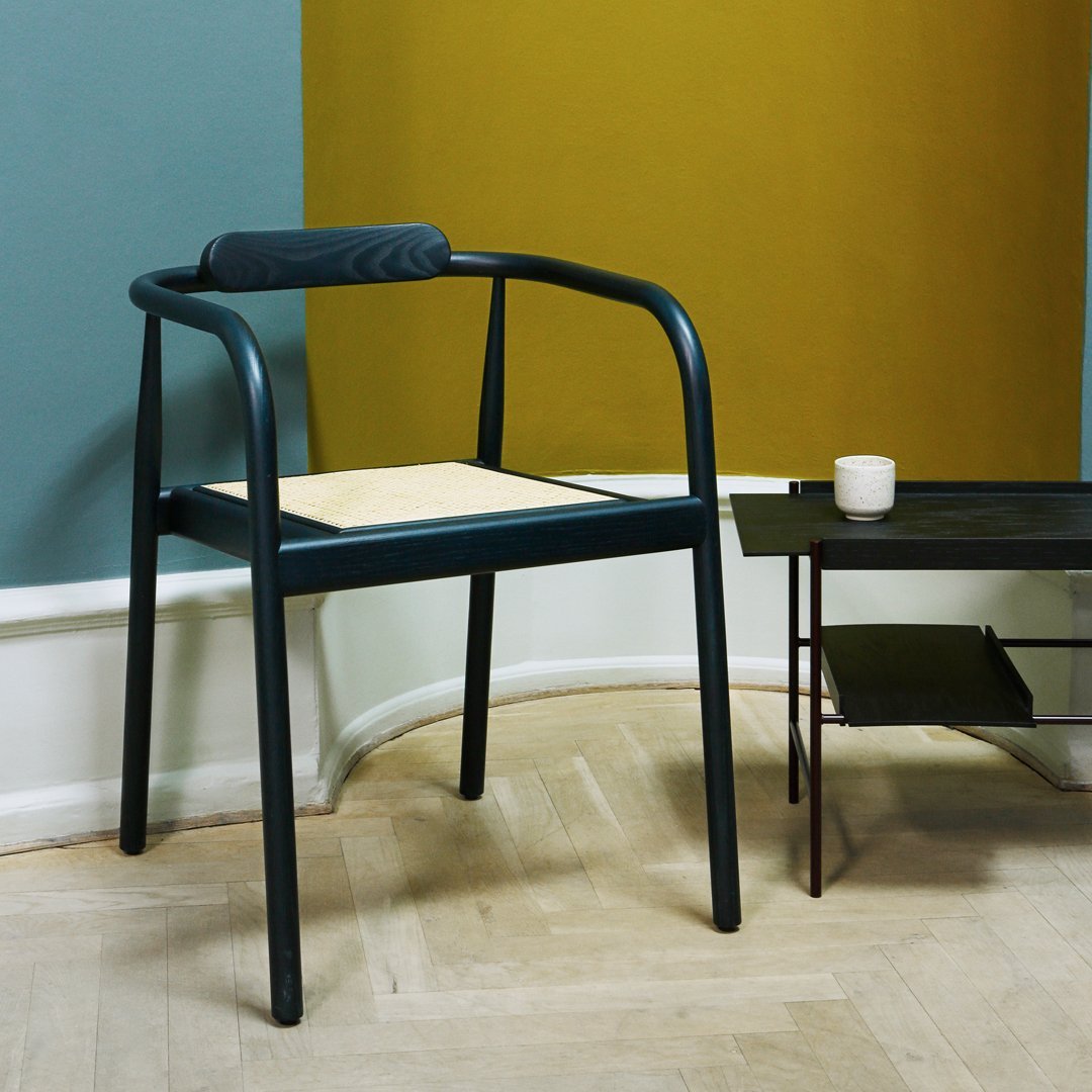Modern blue chair with natural wood seat, set against a muted yellow wall and a stylish black table.