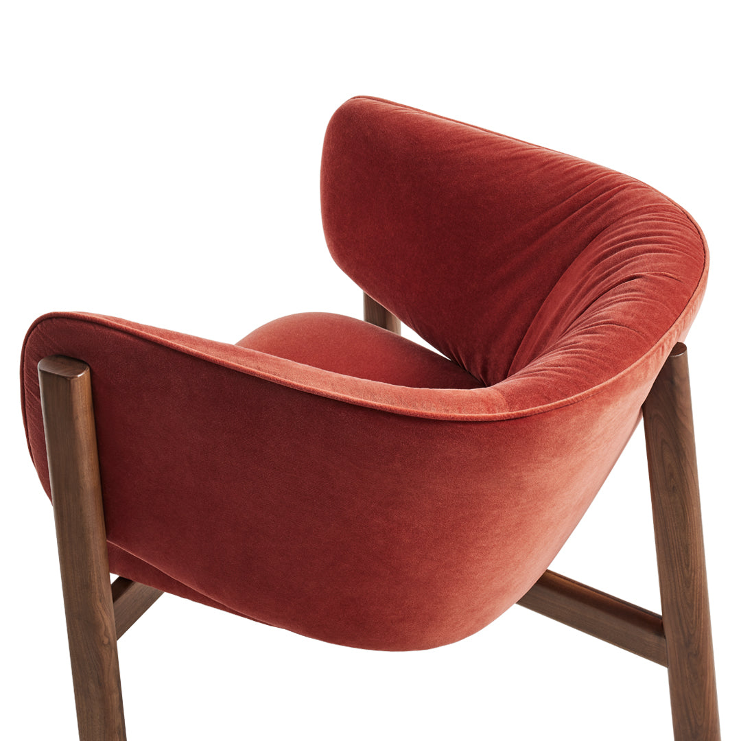 Rust-colored velvet armchair with smooth texture and curved design, featuring wooden legs. Stylish and modern accent piece.