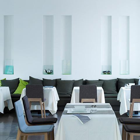 Modern restaurant interior featuring gray seating, white tablecloths, and green accent pillows against light blue walls.