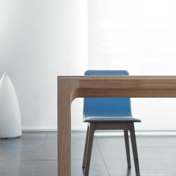 Light wood table edge with smooth curves, featuring a vibrant blue chair against a minimalist backdrop.