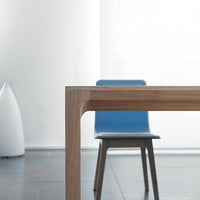 Light wood table edge with smooth curves, featuring a vibrant blue chair against a minimalist backdrop.