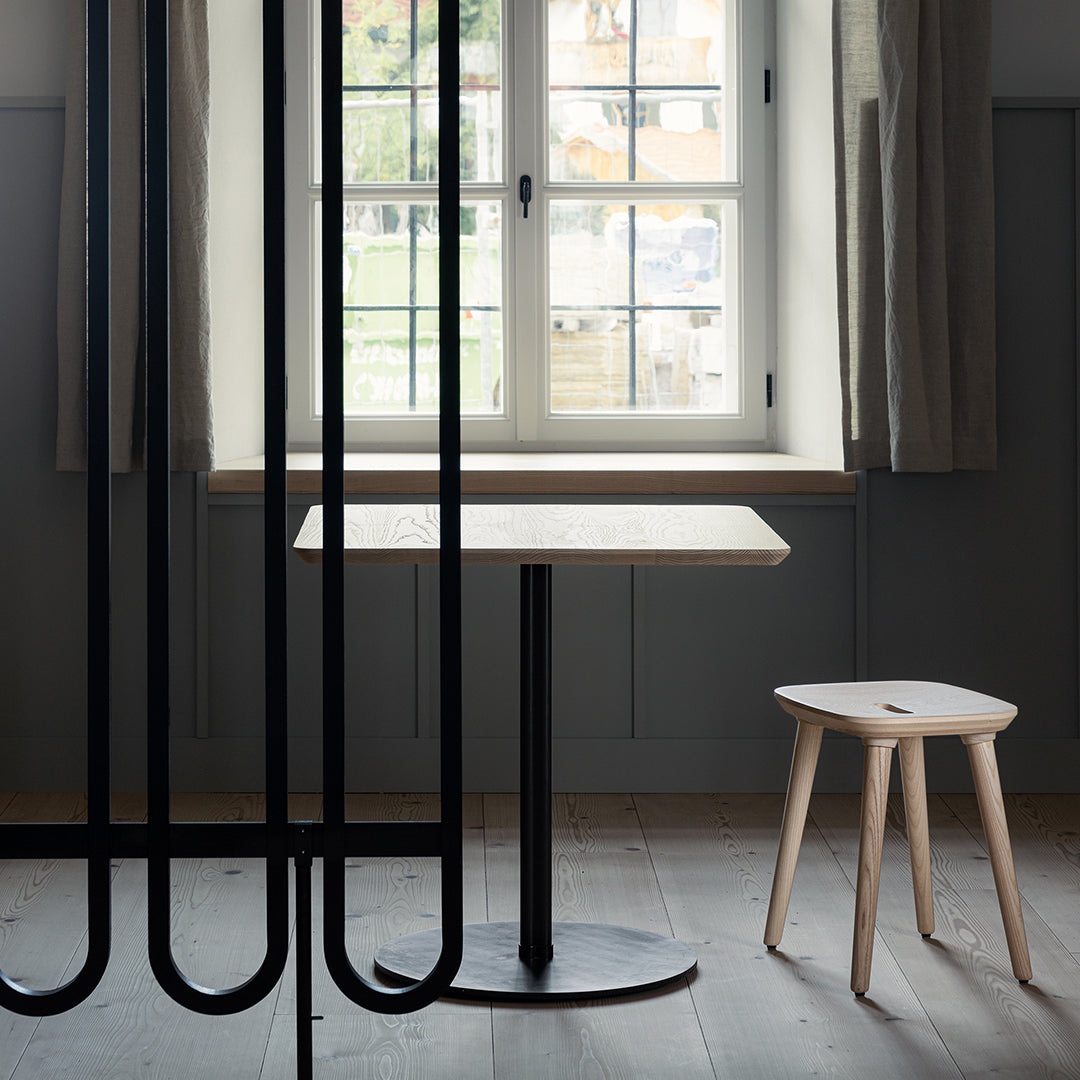 Modern minimalist black and wood table with natural grain pattern, paired with a light wooden stool in a soft, neutral setting.