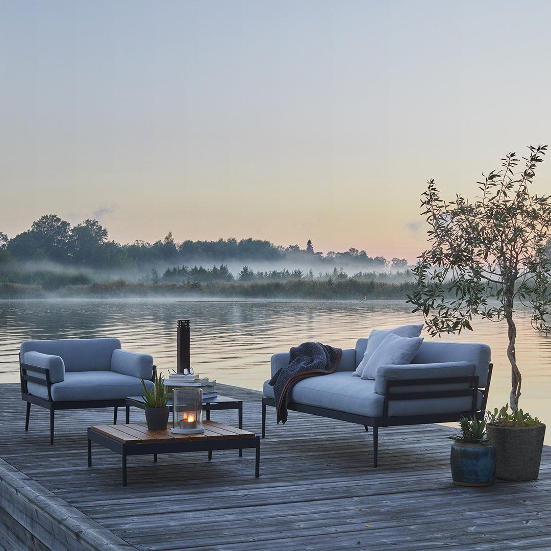 Gray outdoor seating furniture with black metal frames on a wooden deck by a tranquil lake at sunrise.