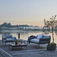 Gray outdoor seating furniture with black metal frames on a wooden deck by a tranquil lake at sunrise.