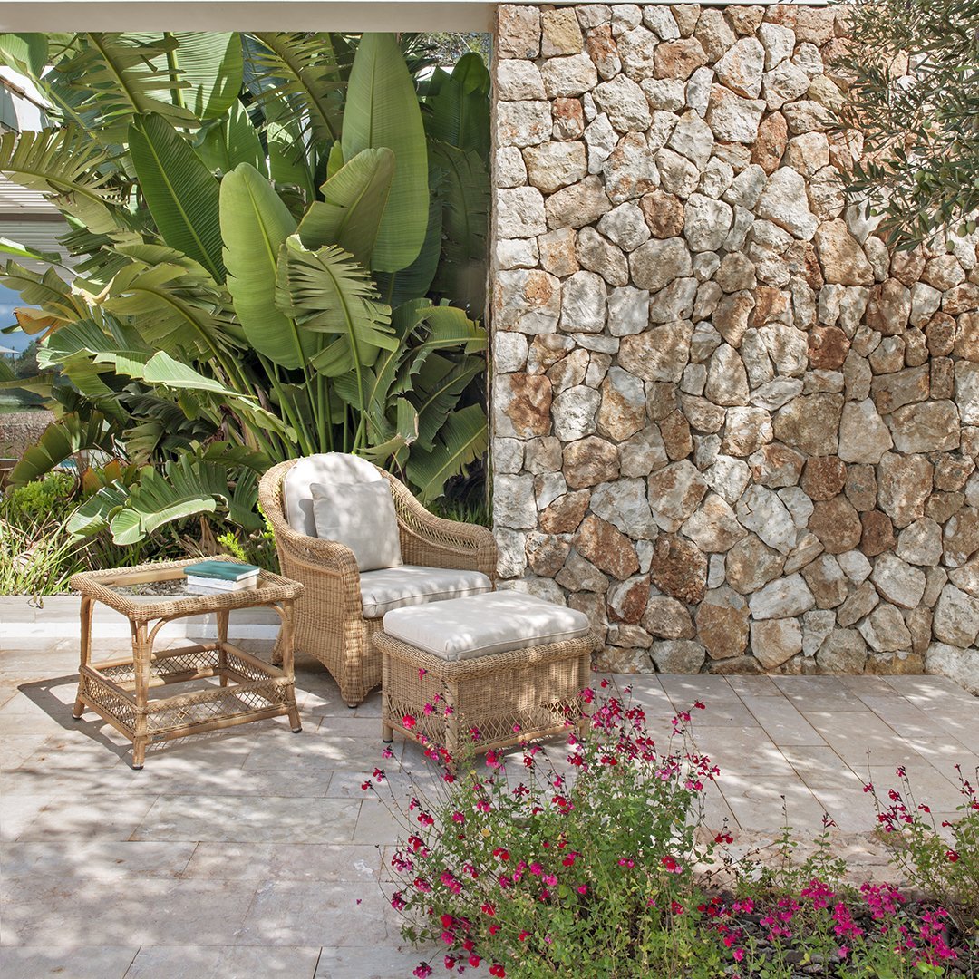 "Natural beige wicker chair with floral cushion, surrounded by lush green plants and a textured stone wall."