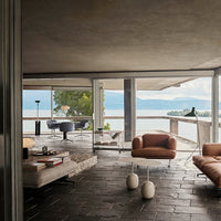 Modern living room with earth-toned furniture, gray stone flooring, and large glass windows showcasing scenic views.