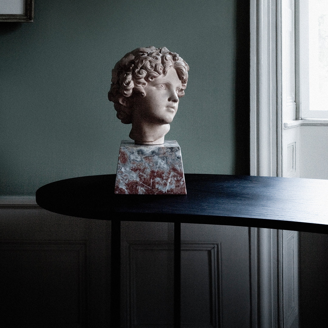 "Artistic marble bust with soft pink tones and intricate reddish patterns on a sleek black table, set against a muted green wall."
