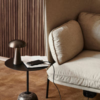 Beige cushion on a sofa next to a sleek black side table with a metallic lamp, set against a textured wood backdrop.