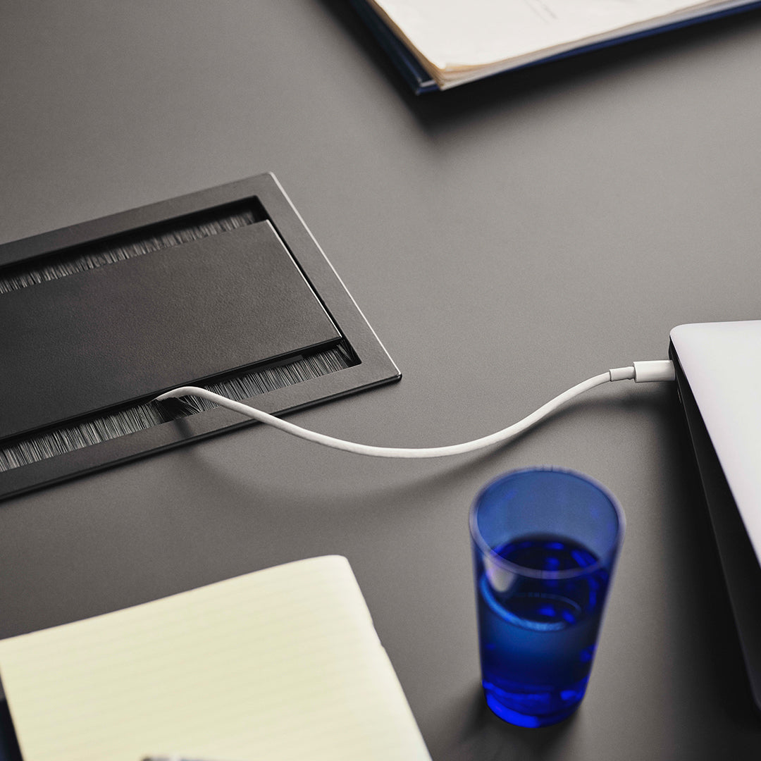 Black cable management box with a gray charging cable, blue glass, and yellow notepad on a dark table.