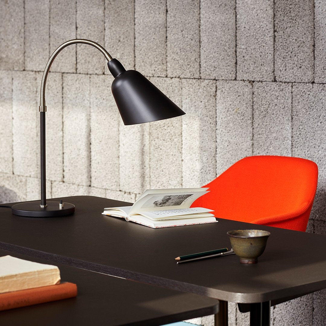 Black desk lamp and bold orange chair on a sleek black table, against textured gray brick wall.