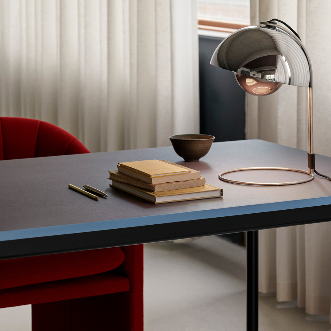 Modern desk with a blue edge, red chair, stacked books, and sleek metallic lamp on a neutral background.