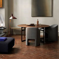 Round wooden dining table with gray upholstered chairs, earthy tones, and a rustic terracotta floor. Modern, minimalist aesthetic.
