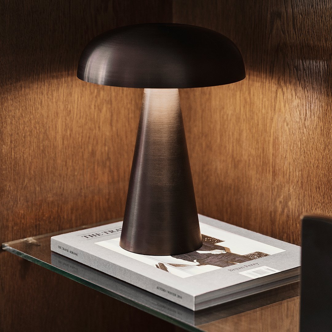 Modern brown metal lamp with a smooth, mushroom-shaped shade on a glass table beside a stylish book.