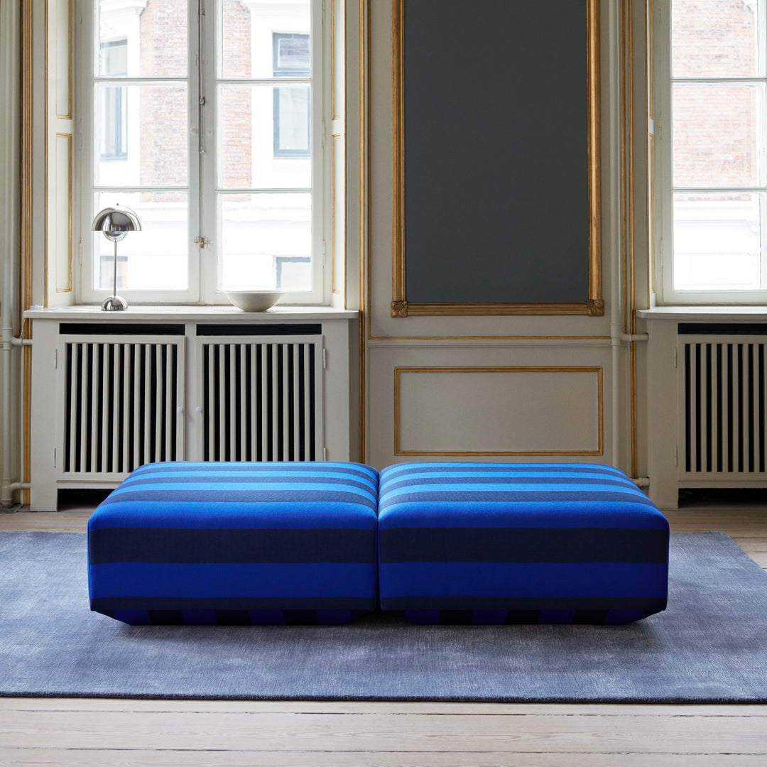 Blue striped pouf with bold horizontal patterns, set in a bright, elegant room with large windows.