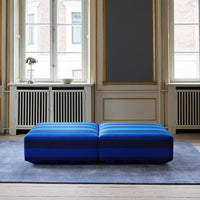 Blue striped ottoman on a soft gray rug in a bright room with large windows and classic decor.