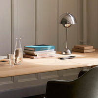 Minimalist desk with natural wood tones, silver lamp, stacked books in muted colors, and glassware. Elegant, modern design.