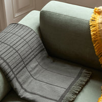 Gray patterned throw blanket with grid design, draped over a green couch, accented by a yellow fringed pillow.