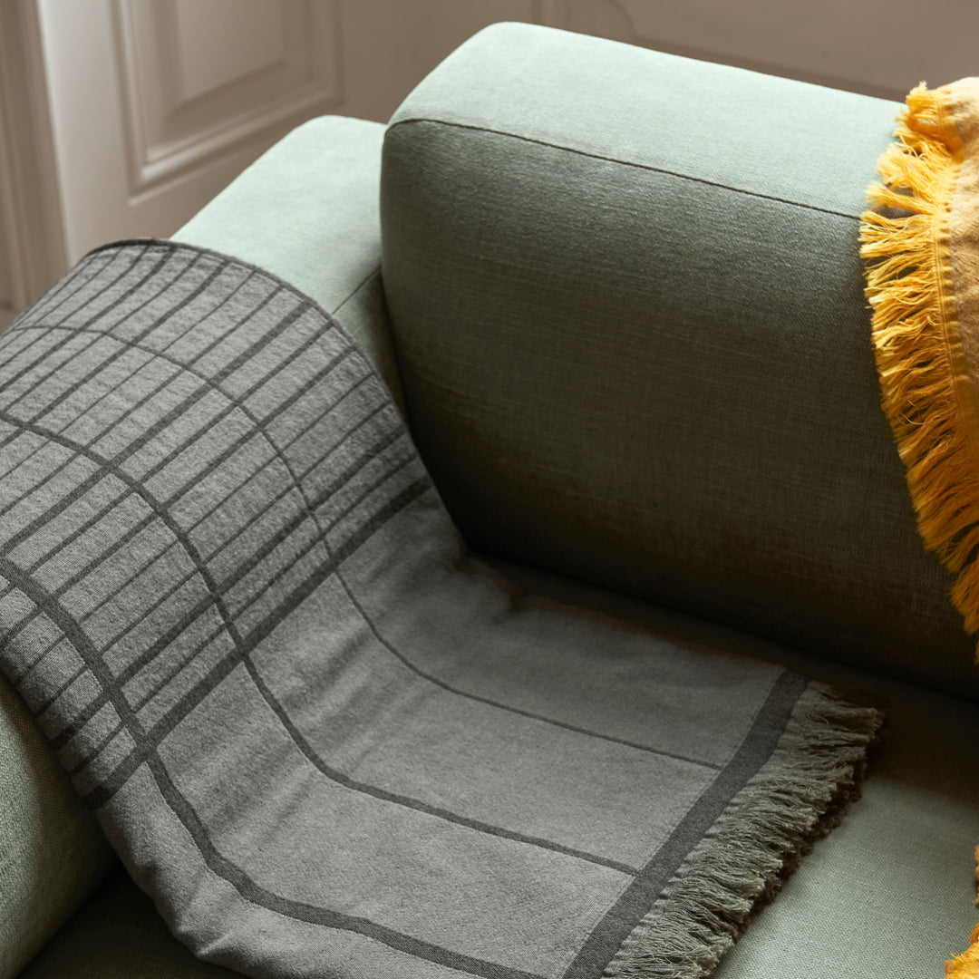 Gray textured blanket with a grid pattern draped over a soft green couch, complemented by a yellow fringed pillow.