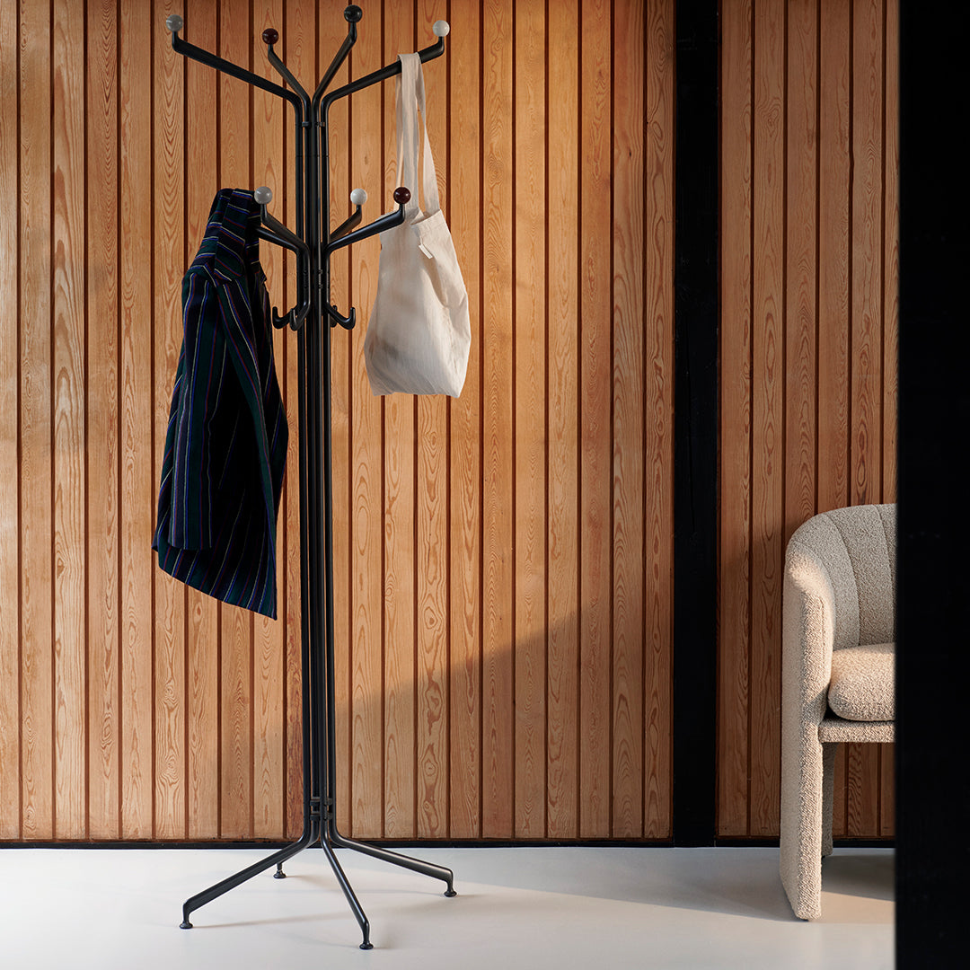 "Black coat rack with a striped navy jacket and white bag against warm wood paneling and a neutral chair."