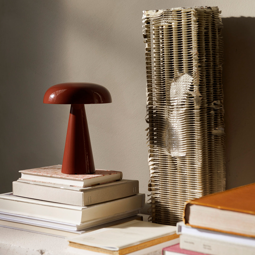 "Deep red mushroom lamp beside a textured beige paper item, stacked on white and pastel-colored books."