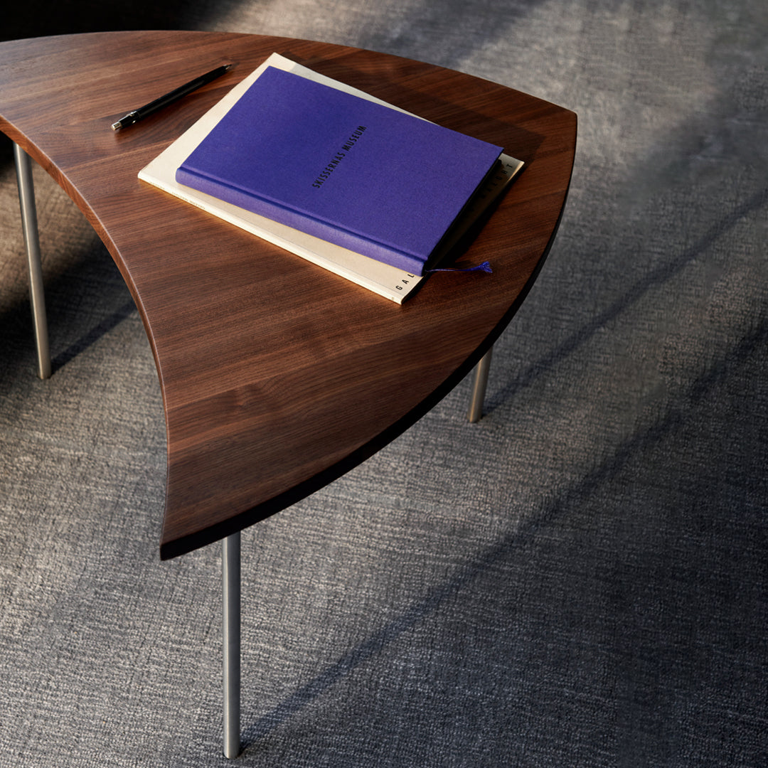 "Curved walnut coffee table with sleek silver legs and a vibrant purple notebook on top against a textured gray background."