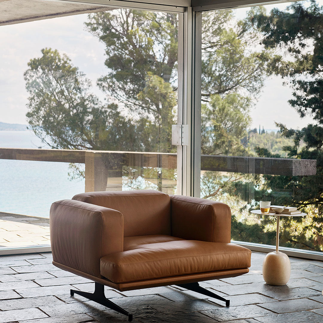 Brown leather armchair with a sleek design, set against a scenic backdrop of greenery and glass walls.