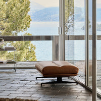 Brown upholstered ottoman on a sleek black metal base, set against a serene outdoor view.