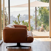 Brown leather swivel chair with a smooth, minimalist design, set against a backdrop of greenery and natural light.