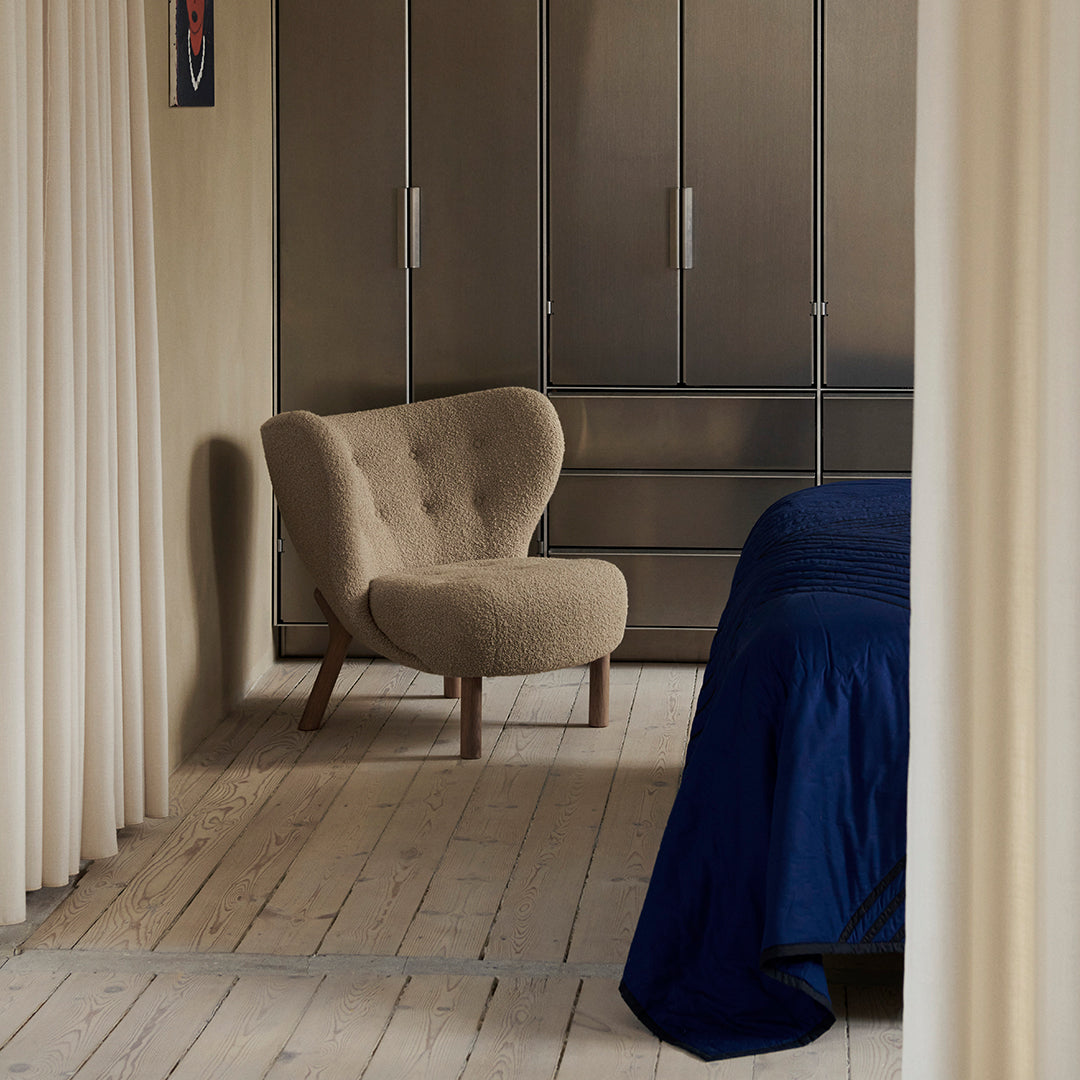 Beige accent chair with rounded back, navy bedding, light wood floor, and soft curtains create a warm, modern interior.