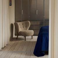 Beige accent chair with rounded back, navy bedding, light wood floor, and soft curtains create a warm, modern interior.