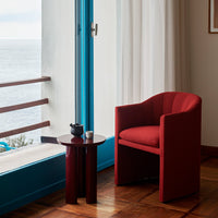 Red upholstered chair next to a round dark table, framed by light curtains and blue window accents.