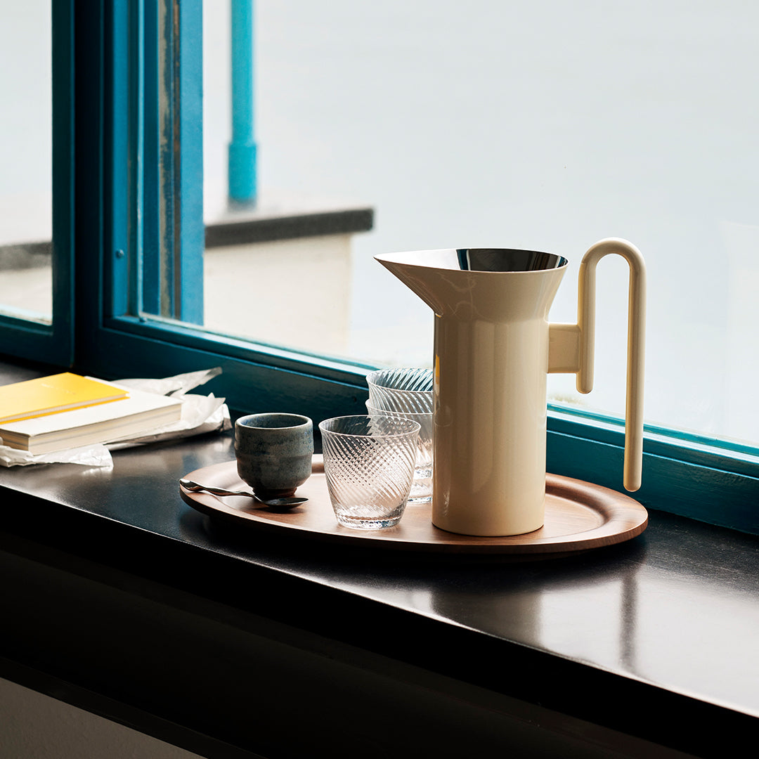 Beige pitcher on a wooden tray, accompanied by clear and frosted glassware, set against a blue window frame.