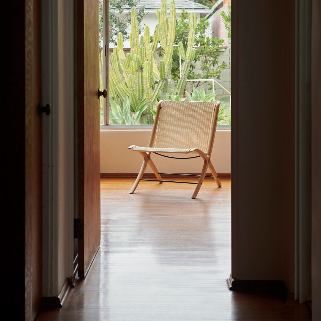 Light wood lounge chair with a woven natural fiber seat, set against a backdrop of green plants and warm wooden floors.