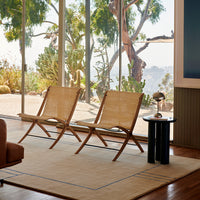 "Two natural rattan chairs on a light beige rug with blue accents, set against a scenic outdoor view."