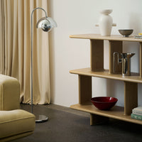 Modern chrome floor lamp beside a light wood shelf; features beige curtains, a white vase, and a dark red bowl.