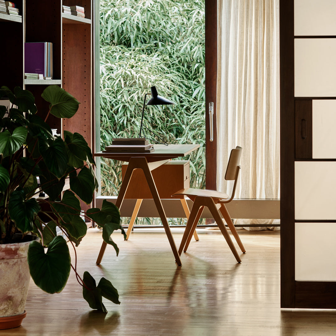 Wooden desk and chair in warm tones, surrounded by greenery and natural light; sleek, modern design with soft textures.