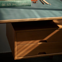 Wooden desk with a light oak finish, teal top, and clean lines, featuring two drawers and warm sunlight shadows.