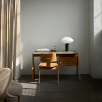"Light wood desk with a modern design, paired with a beige chair and a white lamp, set against a soft gray wall."
