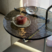 Round glass side table with textured, dark surface; features a pale plate, peach, and clear glass bowl.
