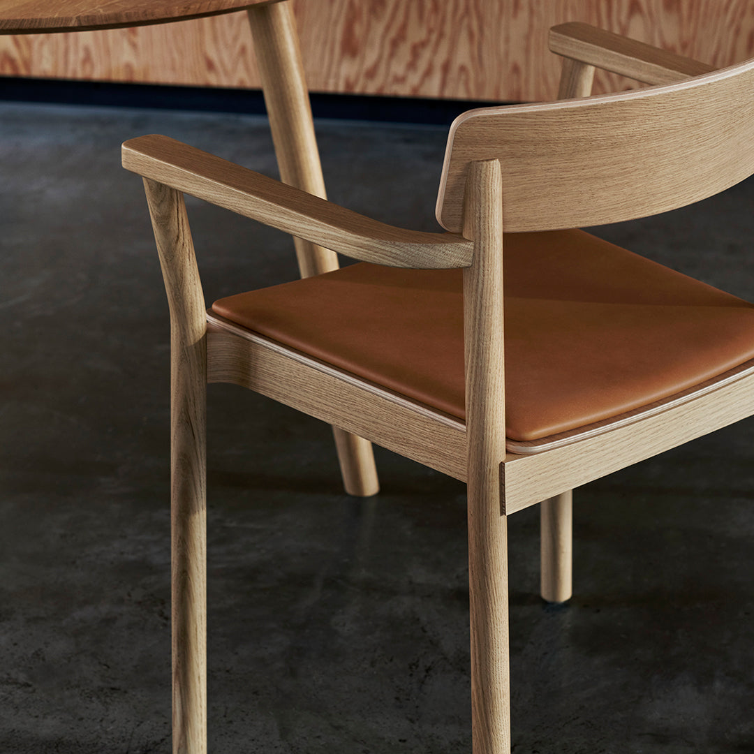 Light wood armchair with a smooth oak finish and a warm brown leather seat, set against a dark floor.