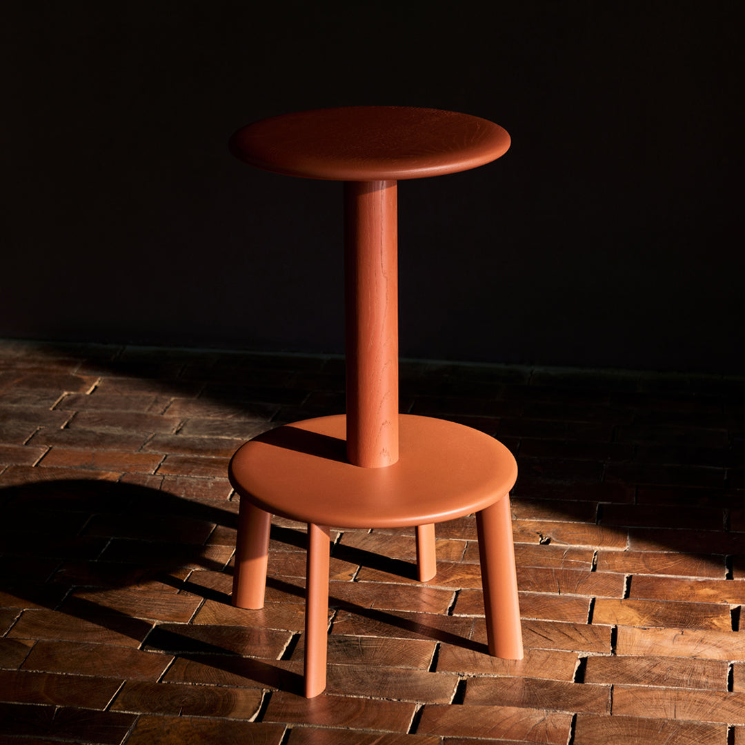 Terra-cotta colored stool with a sleek, minimalist design, featuring solid round seating and a single post structure.
