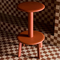 Terracotta two-tiered stool against a checkered brown and beige background. Round, smooth surfaces with a modern design.