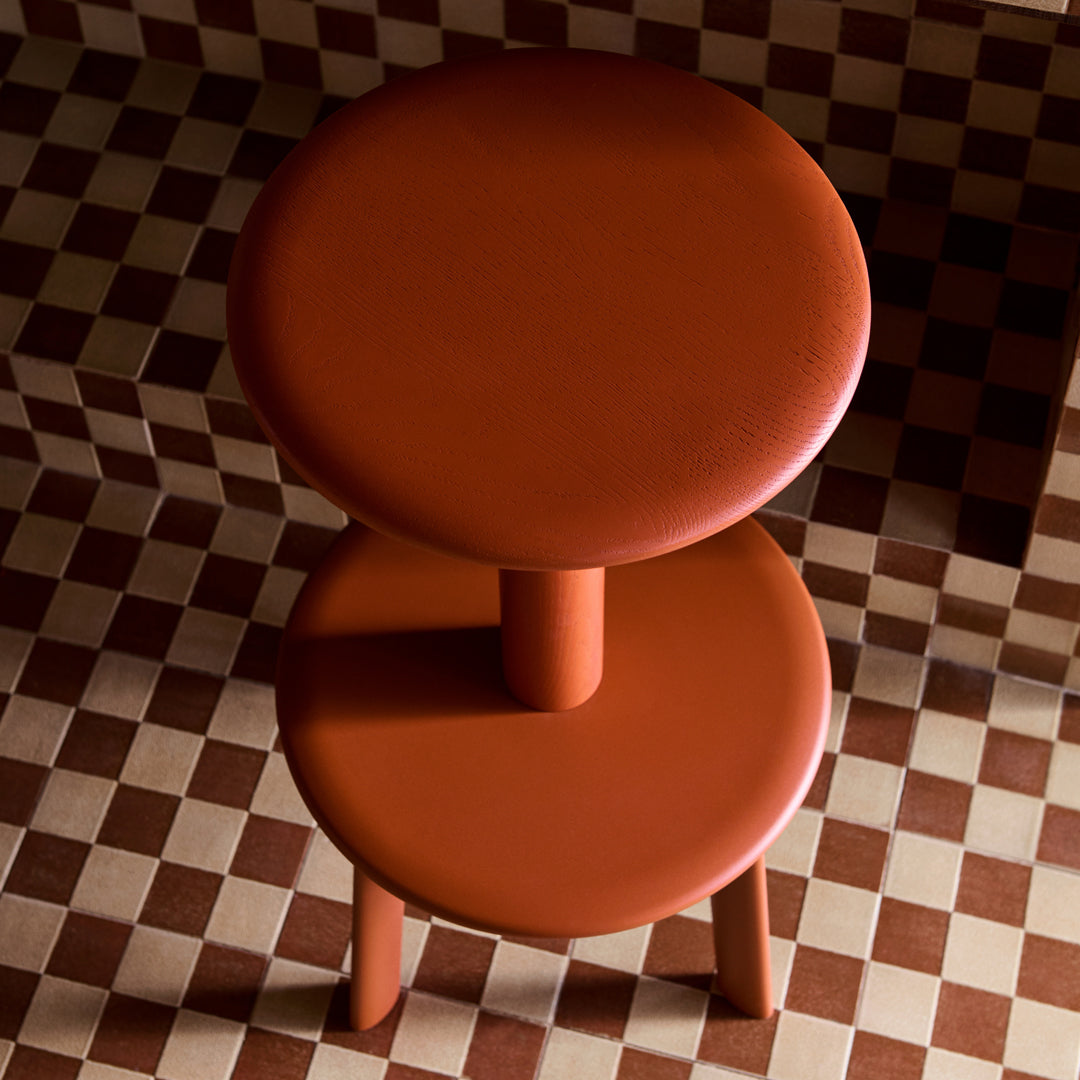Rust-orange cylindrical stool on a checkered brown and cream background, featuring smooth, modern lines.