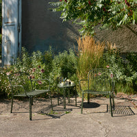 Green metal chairs and table set against lush greenery and beige walls, enhancing outdoor decor with a modern touch.