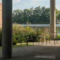 Black and natural wood chairs with minimalist design set against a lush green backdrop near water.