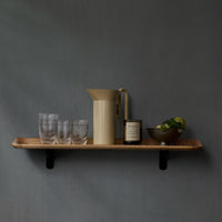 Neutral gray wall with a wooden shelf featuring a beige pitcher, clear glasses, and a dark bowl, accentuated by greenery.