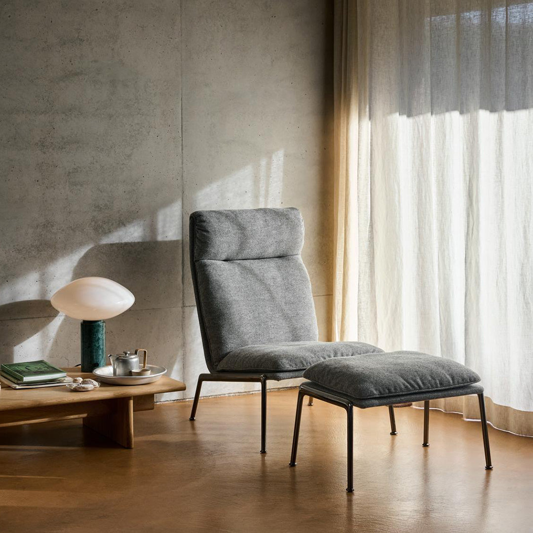 Gray upholstered chair with a matching ottoman, set against a neutral backdrop of light wood and soft, flowing curtains.