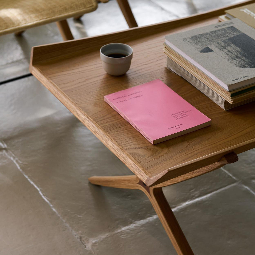 Wooden table with light oak finish, a pink notebook, gray books, and a black cup against a textured stone floor.